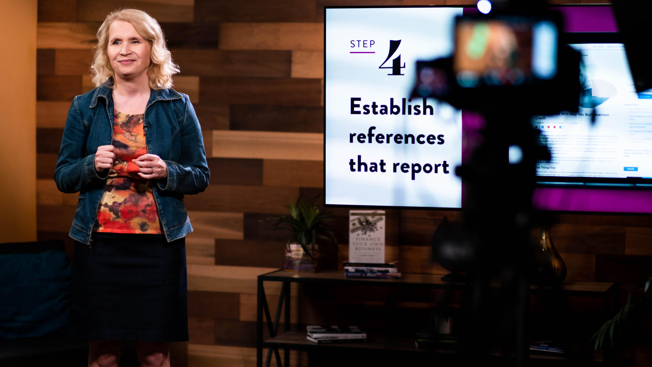 Woman speaking at a conference. There is a news camera and and presentation behind her.